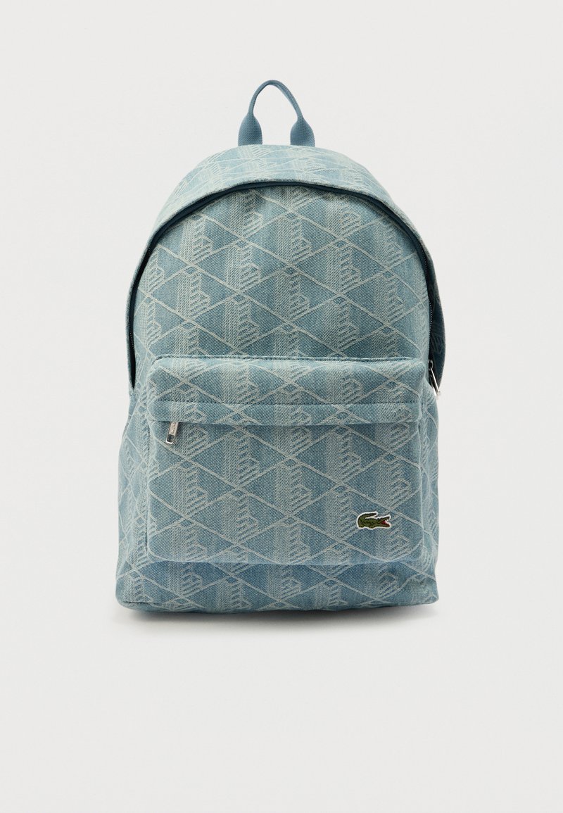 Sac à dos en denim bleu clair avec motif en losange, poche avant zippée et petit logo crocodile vert dans le coin inférieur droit.