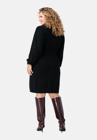 Zwarte gebreide jurk met lange pofmouwen en een ceintuur in de taille, aangevuld met hoogglanzende bordeauxrode kniehoge laarzen.