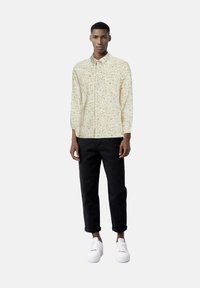 Camicia a bottone con motivo floreale in crema, con accenti marroni e verdi, abbinata a pantaloni neri arrotolati e scarpe da ginnastica bianche.