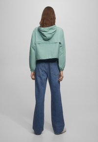 Mintgrön kort jacka med huva och elastiska manschetter, kombinerad med högmidjade blå denimbyxor med utsvängd ben. Slät textur och avslappnad design.