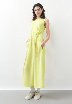 Junge Frau mit lockigem dunklem Haar, die ein ärmelloses hellgrünes Maxikleid mit Taschen und weißen Sneakers trägt, steht vor einer weißen Wand.