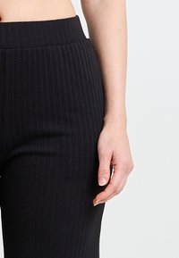 Leggings neri a costine con una vita alta, che mostrano un tessuto testurizzato e un design aderente. Mano appoggiata sulla gamba.