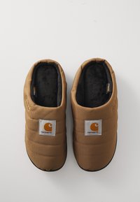 Bruine gewatteerde sloffen met zwarte fleece voering en rubberen zolen. Voorzien van een witte vierkante patch met een oranje logo op elke schoen.