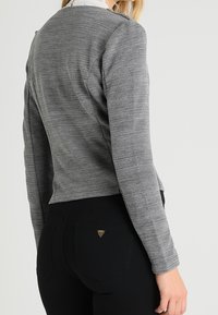 Graues, geripptes Langarmoberteil mit kurzem Schnitt, kombiniert mit schwarzen, hoch taillierten Hosen, die ein kleines metallisches Logo-Detail aufweisen.