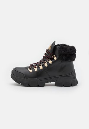 Botte de randonnée en cuir noir avec semelle épaisse, lacets rouges et noirs, œillets dorés et doublure de col en fourrure noire.