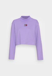 Sudadera recortada de color púrpura con mangas largas, que presenta un cuello alto y un pequeño parche de logotipo de color en la parte frontal. Textura de tela suave.