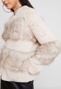 Jas van nepbont in lichtbeige met verschillende texturen, voorzien van lange, zachte mouwen en een cropped model. Geaccentueerd met horizontale strepen.