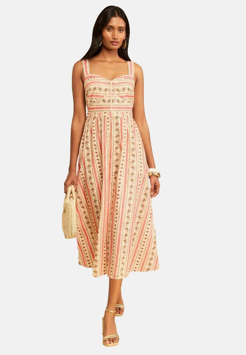 Woman wearing sleeveless, patterned midi dress with vertical stripes, holding woven handbag, with hoop earrings and heeled sandals.