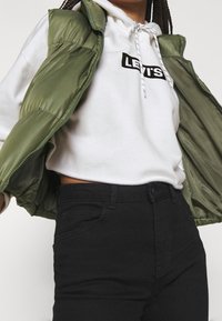 Chaleco acolchado verde caqui sobre sudadera blanca con logotipo negro, combinado con jeans negros de cintura alta. Textura suave; ajuste casual con capucha ajustable.