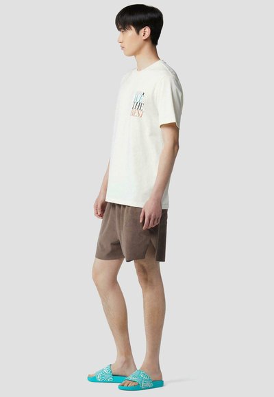 Jeune homme debout de profil portant un T-shirt blanc avec un texte, un short marron et des sandales slides turquoise à motifs sur un fond uni.