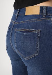 Mörkblå denimjeans med en slät yta, med orange sömmar, två bakfickor och en brun läderetikett vid midjan.