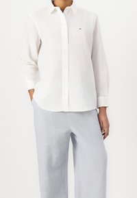 Camisa blanca de manga larga en tejido ligero con cierre de botones en el frente. Incluye un pequeño logo en el pecho, combinada con pantalones gris claro.