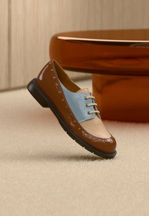 Chaussure marron et bleu clair avec une finition vernie, ornée de clous décoratifs dorés et d'un laçage, posée sur un fond beige texturé.