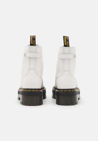 Botas de tobillo de piel blanca con punta redonda, suela de goma negra, costuras amarillas y una lengüeta de tirón con texto en la parte posterior.