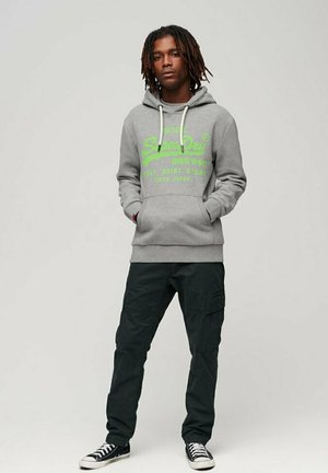 Felpa grigia con logo verde, cordini e tasche frontali, abbinata a pantaloni cargo neri e sneakers nere. Texture liscia.