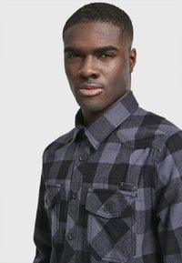 Camisa de franela a cuadros negros y grises con dos bolsillos delanteros, cierre de botones y un parche de contraste en el bolsillo superior izquierdo. Cuello corto.