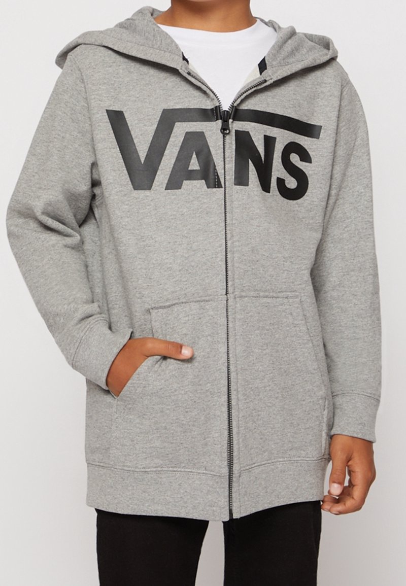 Enfant portant un sweat à capuche gris zippé avec le logo "VANS" en noir, des poches avant et une capuche, assorti à un pantalon noir.