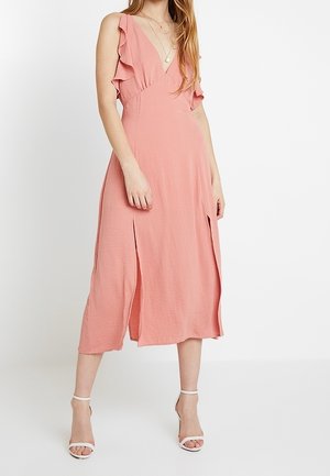 Kobieta w różowej, bezrękawnikowej sukience midi z falbanami na ramiączkach, głębokim dekoltem w kształcie litery V, rozcięciami z przodu i białymi sandałami na kostkę.