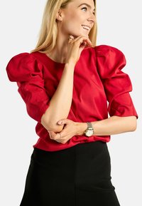 Femeie blondă zâmbitoare, purtând o bluz�ă roșie cu mâneci bufante, fustă neagră și ceas de argint, pozând cu mâinile aproape de față.