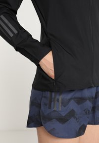 Veste noire à fermeture éclair avec manches longues, comportant trois rayures grises sur le bras gauche. Portée par-dessus un short bleu marine avec un motif en zigzag texturé.