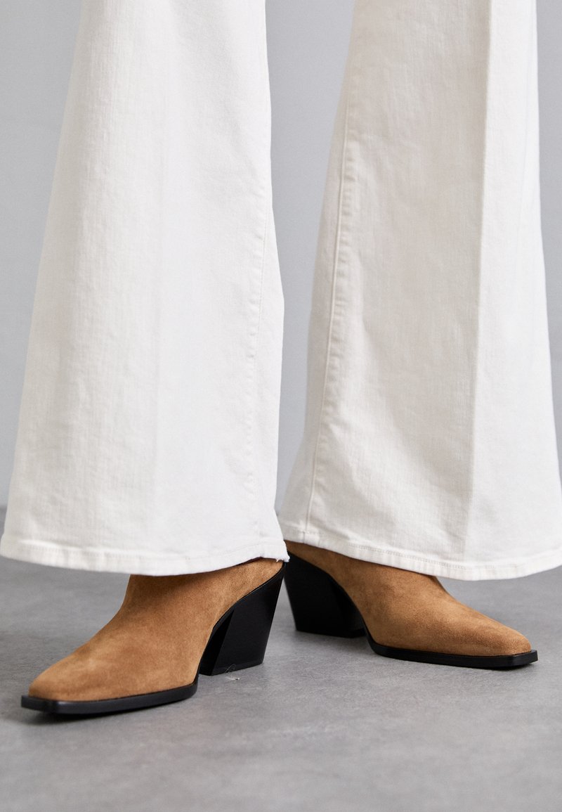 Pruun nahast muulid, millel on terav nina ja must ploki konts, kombineeritud valgete laiade pükstega, hallil pinnal.