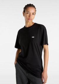 Jeune personne aux cheveux tressés, portant un t-shirt noir Vans et un pantalon sombre, debout avec une main dans la poche sur un fond blanc.