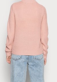 Różowy sweter z teksturowanym splotem, z wysokim kołnierzem i ściągaczem, noszony na jasnoniebieskich, luźnych dżinsach. Prezentuje luźny fason i miękki materiał.