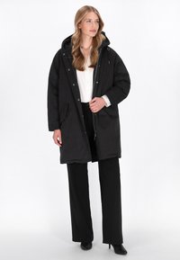 Sort parka med hætte, lynlås og trykknaplukninger, to forlommer og en afslappet pasform, båret over en hvid skjorte og sorte bukser.