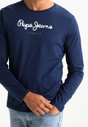 Mand iført en navy langærmet skjorte med hvid "Pepe Jeans London" tekst, kombineret med lyseblå cowboybukser, hånd i lommen.