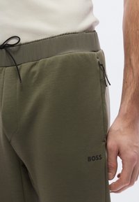 Olivinvihreät juoksuhousut, joissa on kuminauhavyötärö, kiristysnyöri, sivussa vetoketjulliset taskut ja hillitty musta "BOSS"-logo lahkeessa.