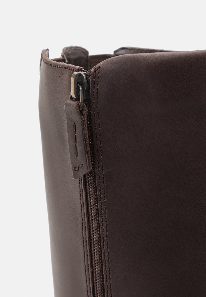 Brązowa skórzana torba z bocznym zamkiem, gładką teksturą i wzmocnionymi szwami na górnej krawędzi z małym skórzanym uchwytem.