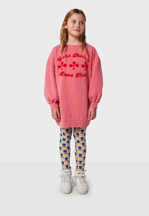 Rosa übergroßer Hoodie mit rotem Text und Grafik, kombiniert mit gemusterten Leggings, die blaue und rote Kreise auf beigem Hintergrund zeigen.