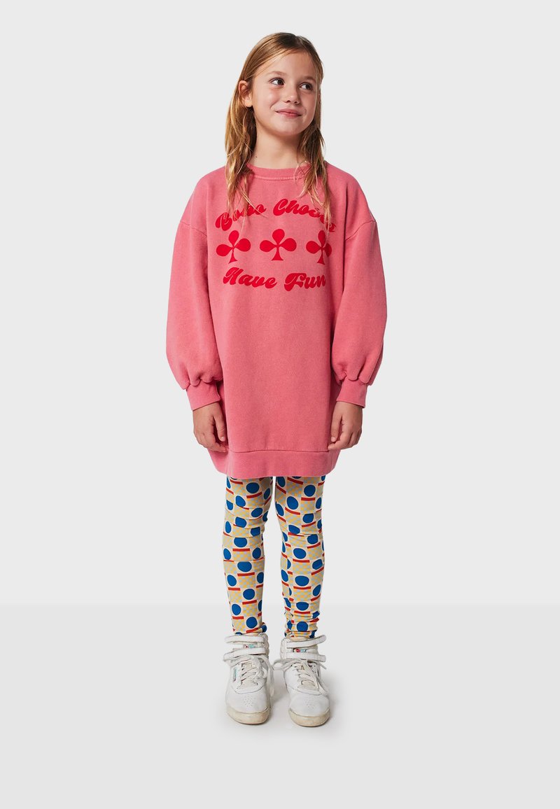 Sweatshirt oversize rosa con testo e grafica rossa, abbinato a leggings stampati con cerchi blu e rossi su uno sfondo beige.