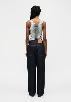 Nainen, jolla on pitkät punotut hiukset ja valkoinen Moschino-hihaton paita sekä korkeavyötäröiset tummansiniset leveälahkeiset farkut, selin päin.