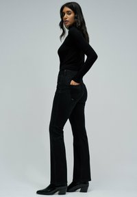 Mujer vestida con un suéter de cuello alto negro, jeans ajustados de corte bootcut negros y botas de tobillo negras, de pie con una mano en el bolsillo trasero contra un fondo liso.