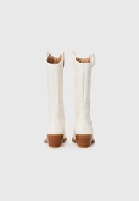 Bottes de cow-boy blanches avec des côtés texturés, un bout pointu et un petit talon en bois. Elles possèdent des pattes de traction en haut pour un enfilage facile.