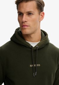 Sudadera con capucha de forro polar de color verde oscuro, con capucha ajustable y un logo crema en el pecho, con una textura suave.