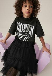 T-shirt graphique noir avec impression "Rolling Stones", associé à une jupe en tulle noir superposée. L'enfant est assis sur une chaise violet clair.