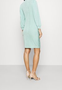 Robe vert menthe clair avec de longues manches bouffantes, un corsage ajusté et une longueur au genou. Associée à des sandales à talons hauts nude. Tissu texturé.