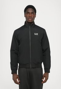 Black bomber jacket featuring a zip front, high collar, elastic cuffs, and hem. Includes a small EA7 logo on the left chest. Smooth texture.