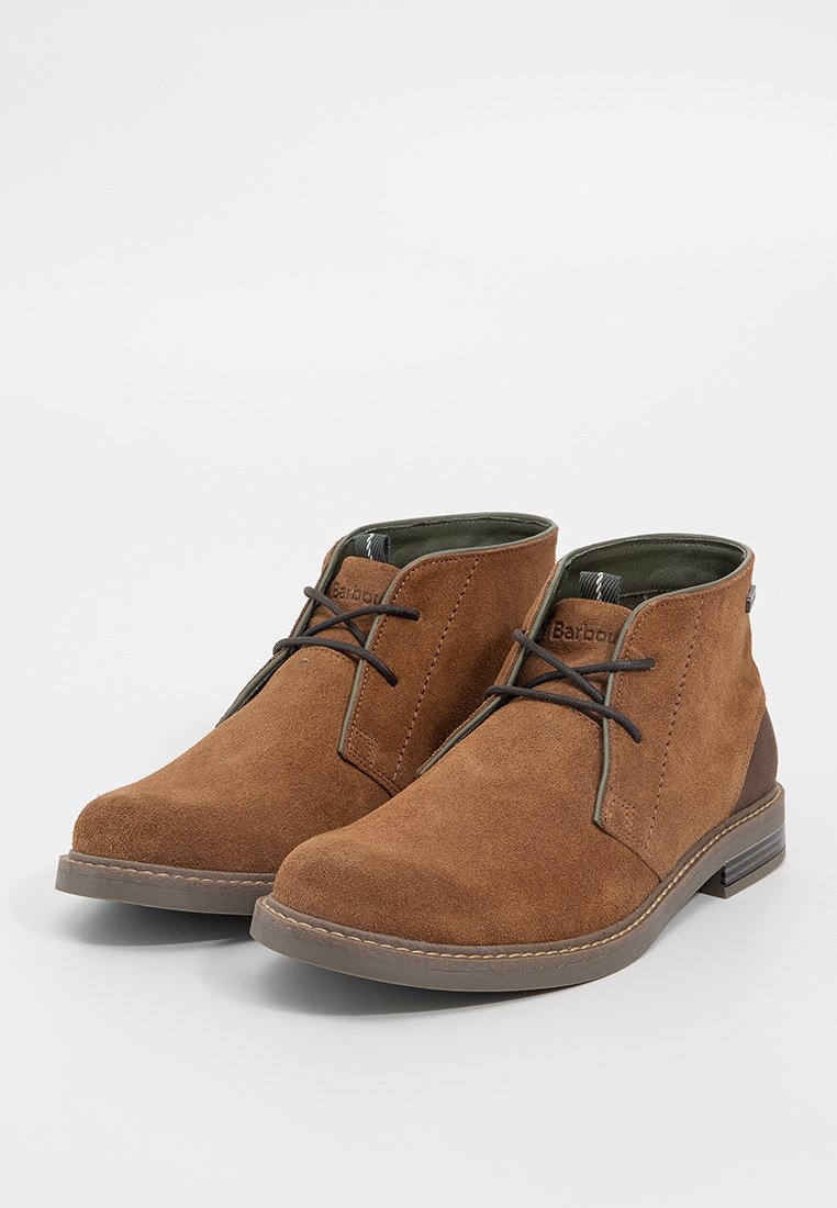 Stivaletti in suede marrone con punta arrotondata, lacci minimal e finitura testurizzata. Presentano un rivestimento verde scuro a contrasto e suola in gomma.