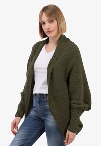 Junge Frau, die ein weißes T-Shirt, blaue Jeans und eine übergroße olivgrüne Strickjacke mit Rauten-Muster trägt.