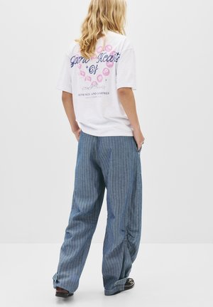 T-shirt graphique blanc avec le texte "Cœurs de jardin", associé à un pantalon large rayé bleu avec des poches latérales et une coupe décontractée.