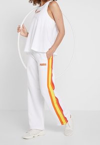 Top blanco sin mangas con dobladillo redondeado y logo; pantalones blancos con franjas laterales rojas y amarillas; combinados con zapatos claros.