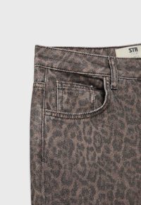 Detailní záběr hnědých džín s leopardím potiskem, ukazující přední kapsu, poutko na opasek a částečný pas s štítkem.