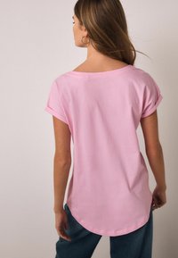 Tricou roz din bumbac cu mâneci suflecate, textură moale, tiv rotunjit și croială lejeră, purtat cu pantaloni din denim albastru. Vedere din spate.