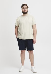 Beige bomulls T-skjorte med korte ermer, kombinert med marineblå shorts. Modellen har på seg hvite joggesko. Enkel, uformell design uten synlige mønstre.