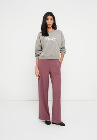 Γκρι φούτερ με το κείμενο «Smile», σε συνδυασμό με φαρδιά βυσσινί παντελόνια. Το μοντέλο στέκεται με τα χέρια στις τσέπες, φορώντας μαύρα μπαλαρίνες.