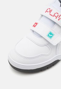 Scarpa da atleta bianca con cinturini in velcro, caratterizzata da un'etichetta rossa "PLAY", un segno di spunta blu e una croce rossa sui cinturini. Suola nera.