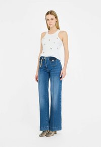 Top sin mangas blanco con adornos de tela, combinado con jeans de pierna ancha azules que cuentan con bolsillos frontales y herrajes en tono dorado.